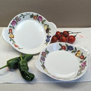Può includere: Due piatti da portata in ceramica bianca con disegni floreali e fruttati sui bordi. I piatti hanno manici e sono circondati da verdure fresche, tra cui peperoni verdi e pomodori.