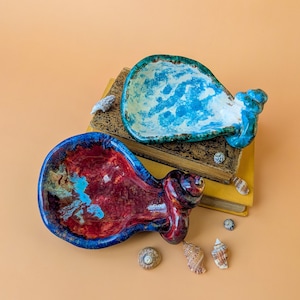 Könnte beinhalten: Zwei Keramikschalen in Muschelform, eine mit einer leuchtend blau-weißen Sprenkelglasur und die andere mit einer tiefroten und blauen Marmorierung. Beide Stücke haben eine glänzende Textur und ein Spiraldetail am schmalen Ende der Muschel.