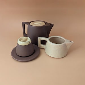 Può includere: Una teiera in ceramica marrone scuro con coperchio abbinato, una zuccheriera marrone scuro con coperchio su piattino abbinato e una lattiera in ceramica beige chiaro. Ogni pezzo presenta un manico quadrato e un design semplice e funzionale.