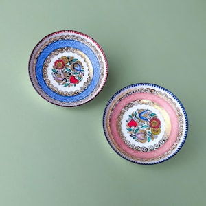 Può includere: Due piattini rotondi in ceramica con intricati disegni dipinti a mano. Un piattino presenta un bordo blu a tratteggio incrociato, l'altro un bordo rosa a tratteggio incrociato. Entrambi mostrano un motivo floreale centrale con cuori rossi e oro.