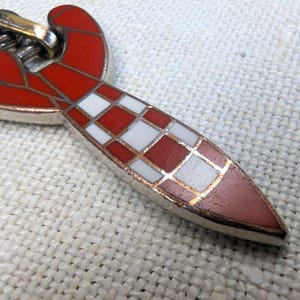 May include: Close-up of a silver-toned pendant with a red and white checkered design. The pendant has a red, curved top and a pointed, checkered bottom. The checkered pattern is red and white with gold-colored accents.