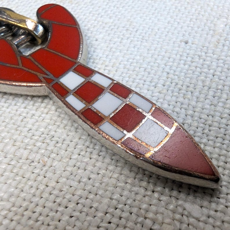 May include: Close-up of a silver-toned pendant with a red and white checkered design. The pendant has a red, curved top and a pointed, checkered bottom. The checkered pattern is red and white with gold-colored accents.