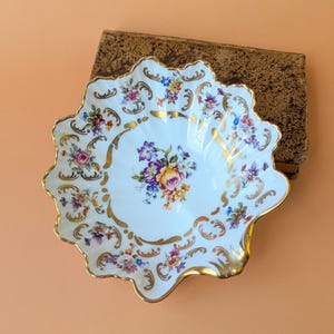 Könnte beinhalten: Eine weiße Porzellanschale mit gewelltem Rand und einem zentralen Blumenstrauß in Rosa, Gelb und Violett. Der Rand ist mit goldenen Schnörkeln und kleineren Blumenmotiven verziert, wobei die gesamte Kante mit Gold bemalt ist.