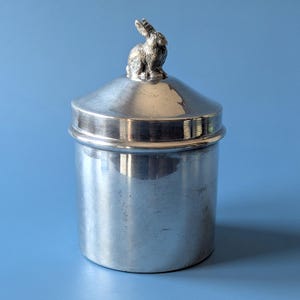 Könnte beinhalten: Ein zylindrischer Behälter aus silberfarbenem Metall mit einer glatten, reflektierenden Oberfläche. Der Deckel ist in der Mitte mit einer kleinen, detaillierten Hasenfigur versehen, die als Griff für den abnehmbaren Deckel dient.