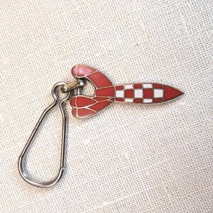 May include: A red and white rocket-shaped keychain with a silver clasp. The rocket has a checkered pattern on the body and red fins. The keychain is made of metal and is likely enamel.