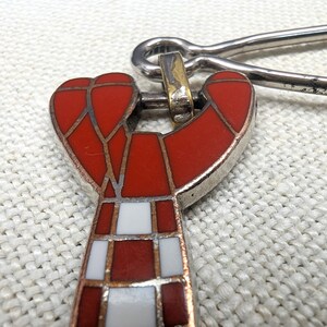 May include: Close-up of a red and white enamel pendant with a silver-tone metal clasp. The pendant features a geometric design with red and white squares and a silver-tone metal border. The clasp is a simple pin.