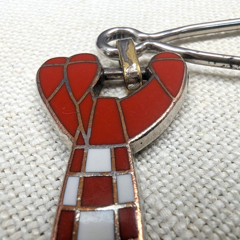 May include: Close-up of a red and white enamel pendant with a silver-tone metal clasp. The pendant features a geometric design with red and white squares and a silver-tone metal border. The clasp is a simple pin.