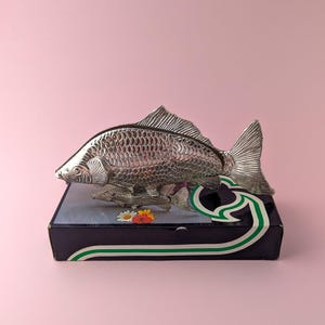 May include: A silver-toned metal fish figurine with detailed scale textures, positioned atop a rectangular dark blue box. The box features a graphic print of a smaller fish and a cluster of white, yellow, and red flowers, accented by a green and white curved line.