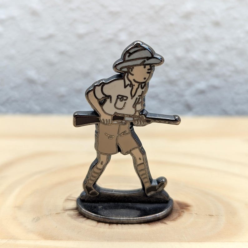 May include: A metal figurine depicting a person in a safari outfit, holding a rifle. The figure is in a walking pose, wearing a hat and shorts. The color palette includes silver, beige, and brown tones. The figurine is on a circular base.