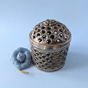 Op de afbeelding: Een zilverkleurige metalen houder met een afneembaar deksel voorzien van een uitgesneden schubpatroon en een kleine gebeeldhouwde bloem bovenop. Naast de houder ligt een enkele lichtblauwe stoffen bloem met een dunne gele steel aan de basis.