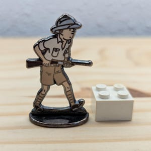 May include: A metal figurine of a person in safari attire, holding a rifle, with a white hat and shorts. The figurine is on a dark base, next to a white Lego brick. The figure is approximately 3.8 cm tall.