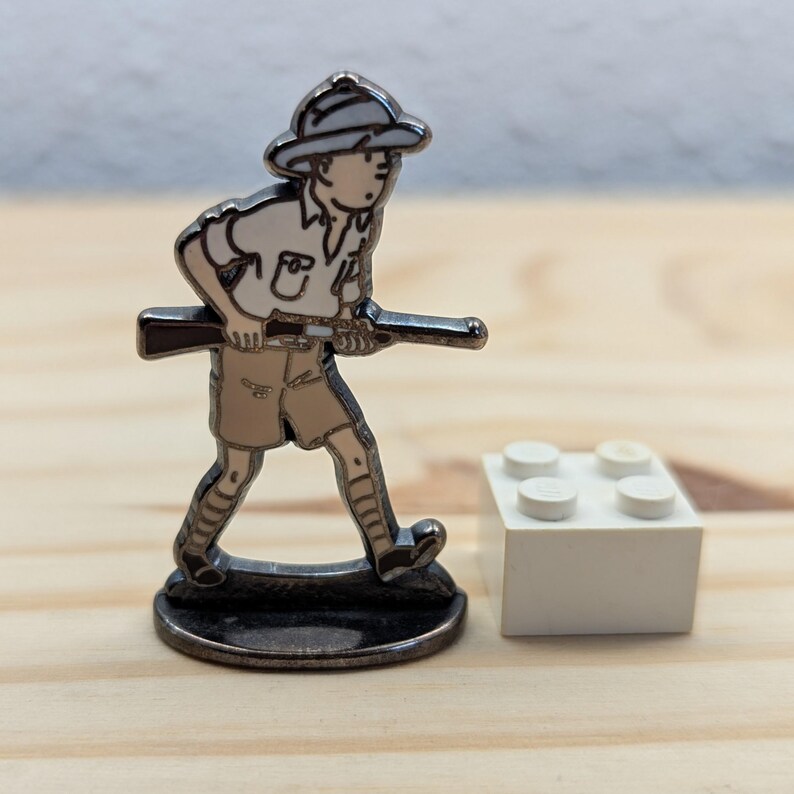 May include: A metal figurine of a person in safari attire, holding a rifle, with a white hat and shorts. The figurine is on a dark base, next to a white Lego brick. The figure is approximately 3.8 cm tall.