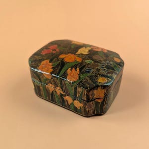 Op de afbeelding: Een rechthoekige zwarte doos met afgeronde hoeken en een handgeschilderd bloemmotief. Het oppervlak toont diverse oranje, gele, rode en blauwe bloemen met groene stengels en bladeren tegen een donkere achtergrond met fijne gouden lijnen.