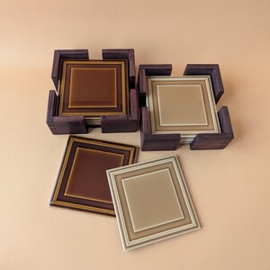 May include: Two dark brown wooden holders containing stacks of square ceramic coasters. One set features deep chocolate brown tiles with lighter brown borders, while the other set contains light beige tiles with matching beige borders.