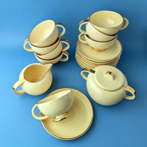 Op de afbeelding: Een crèmekleurige keramische theeset bestaande uit meerdere kopjes met oren, bijpassende schoteltjes, een melkkannetje en een suikerpot met deksel. Elk onderdeel is afgewerkt met een dunne gouden rand langs de randen voor een uniforme uitstraling.