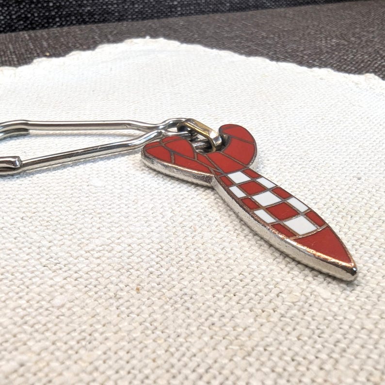 May include: A red and white patterned key chain with a silver-colored clasp. The key chain has a surfboard shape with a red and white checkered design. The key chain is made of metal and has a shiny finish.