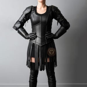 Pode incluir: Conjunto de armadura de couro preto com couraça, ombreiras, protetores de braço, luvas e uma saia com tiras verticais. A armadura é adornada com rebites e fivelas. A modelo usa botas pretas e um top preto.