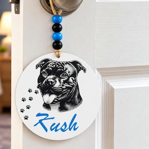 May include: A round white wooden door sign hanging from a silver doorknob by a jute rope with alternating blue and black beads. The sign features a black and white illustration of a dog, small black paw prints, and the blue text Kush.
