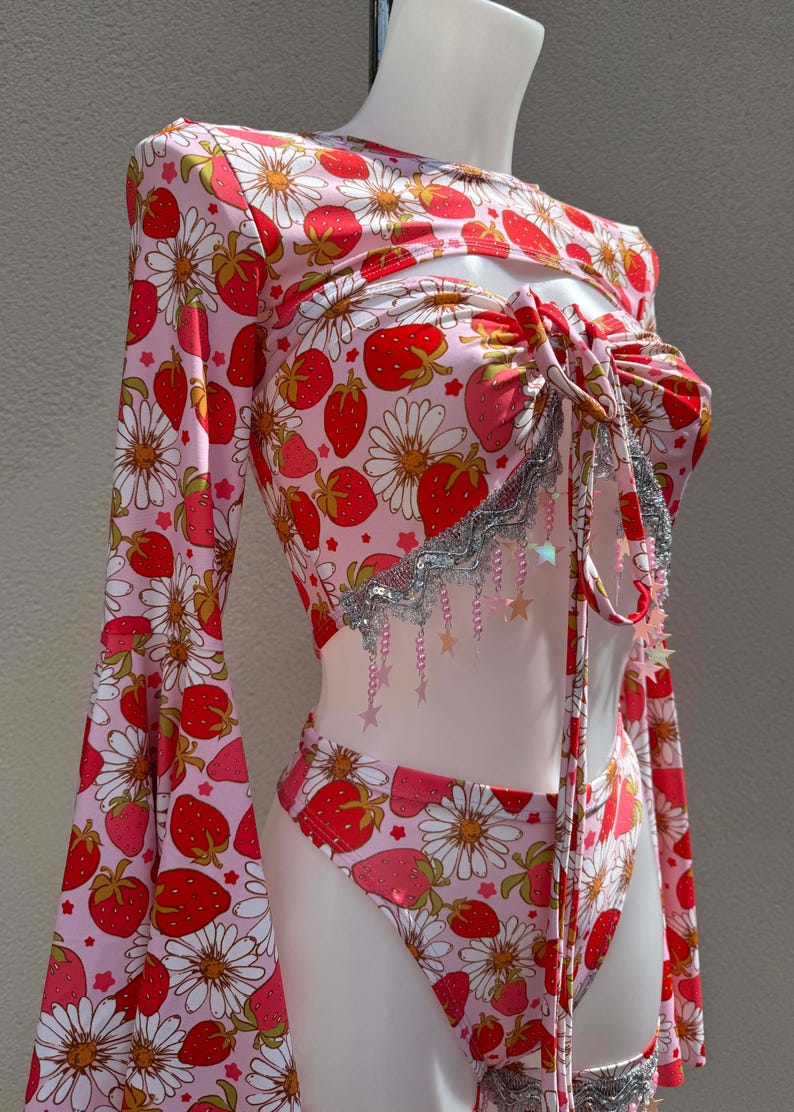 May include: A pink two-piece outfit with a strawberry and daisy print. The top has long bell sleeves and a cut-out design. The outfit is adorned with silver trim and star-shaped embellishments, creating a playful and feminine aesthetic.