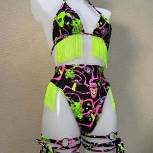 May include: Black bikini set with neon green fringe and high-waisted bottoms. The fabric features a repeating pattern of green aliens, pink eyes, and space-themed graphics. Includes leg straps.