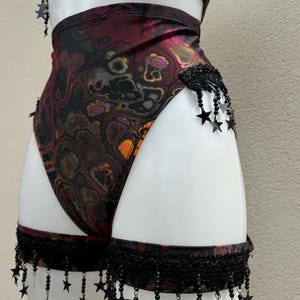 May include: A one-piece swimsuit with a swirling, abstract pattern in shades of burgundy, orange, and black. The swimsuit is adorned with black star-shaped embellishments and fringe detailing. The garment is displayed on a white mannequin.