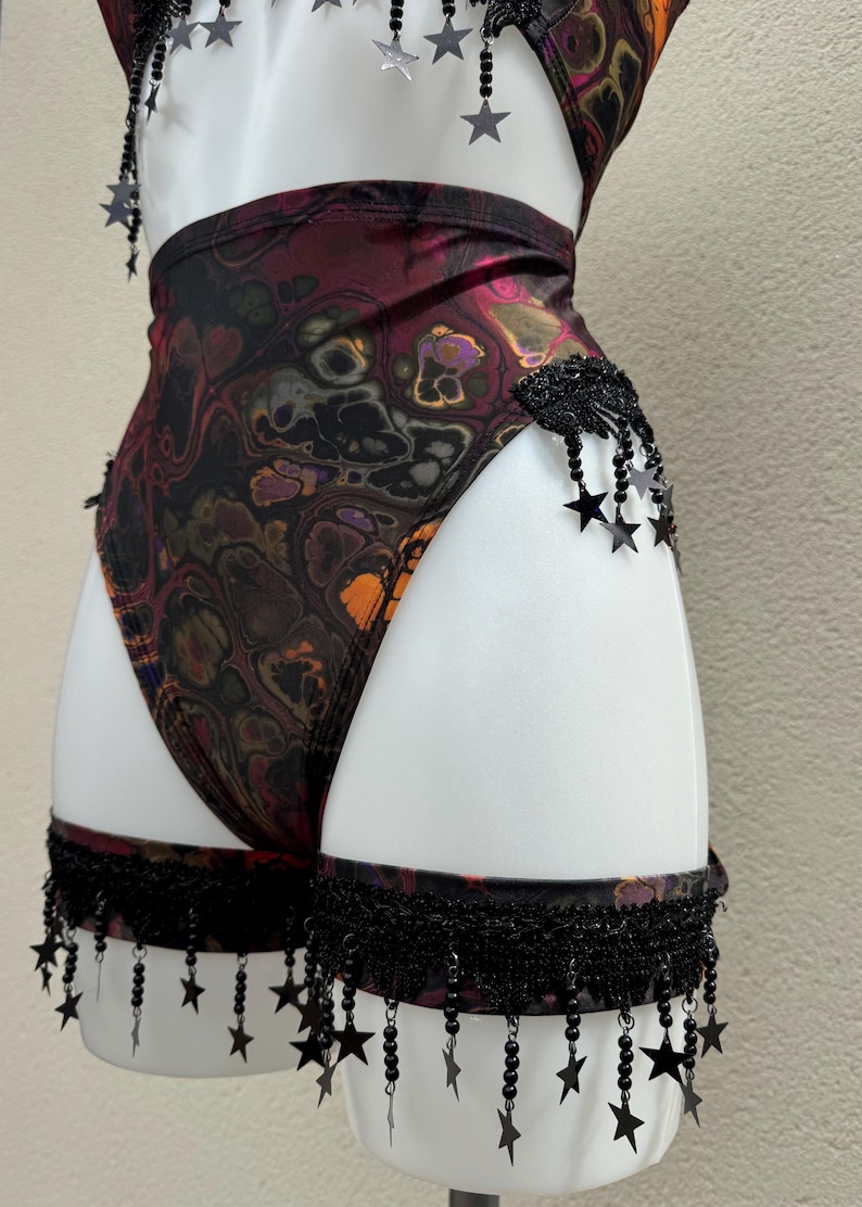 May include: A one-piece swimsuit with a swirling, abstract pattern in shades of burgundy, orange, and black. The swimsuit is adorned with black star-shaped embellishments and fringe detailing. The garment is displayed on a white mannequin.
