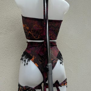 May include: A two-piece outfit, comprising a halter top and high-waisted bottoms, displayed on a mannequin. The set has a swirling burgundy, orange, and black pattern. Black fringe with star accents decorates the bottoms. The outfit is tied with bows.