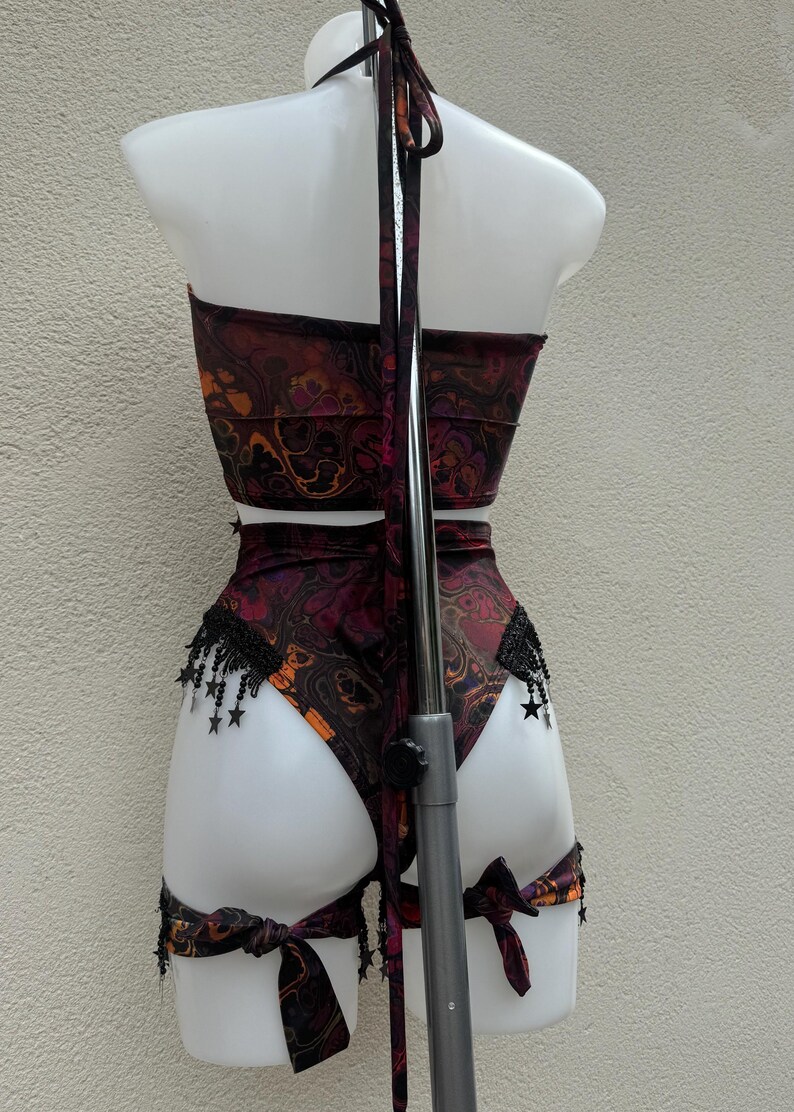 May include: A two-piece outfit, comprising a halter top and high-waisted bottoms, displayed on a mannequin. The set has a swirling burgundy, orange, and black pattern. Black fringe with star accents decorates the bottoms. The outfit is tied with bows.