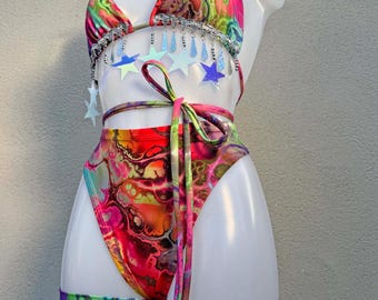 May include: A two-piece bikini set featuring a vibrant, multi-colored fluid marble print in shades of pink, orange, green, and blue. The top and leg garter accessories are adorned with silver-toned trim, dangling metallic beads, and star-shaped charms.