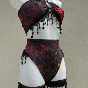 May include: A two-piece outfit featuring a halter top and high-waisted bottoms. The fabric displays a swirling pattern in red, black, and orange. Black star-shaped fringe decorates the top and bottom edges, adding a decorative element.