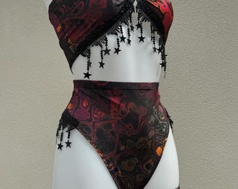 May include: A two-piece outfit with a halter top and high-waisted bottoms. The fabric has a swirling pattern in shades of red, black, and orange. Black star-shaped fringe adorns the top and bottom edges, adding a decorative element.