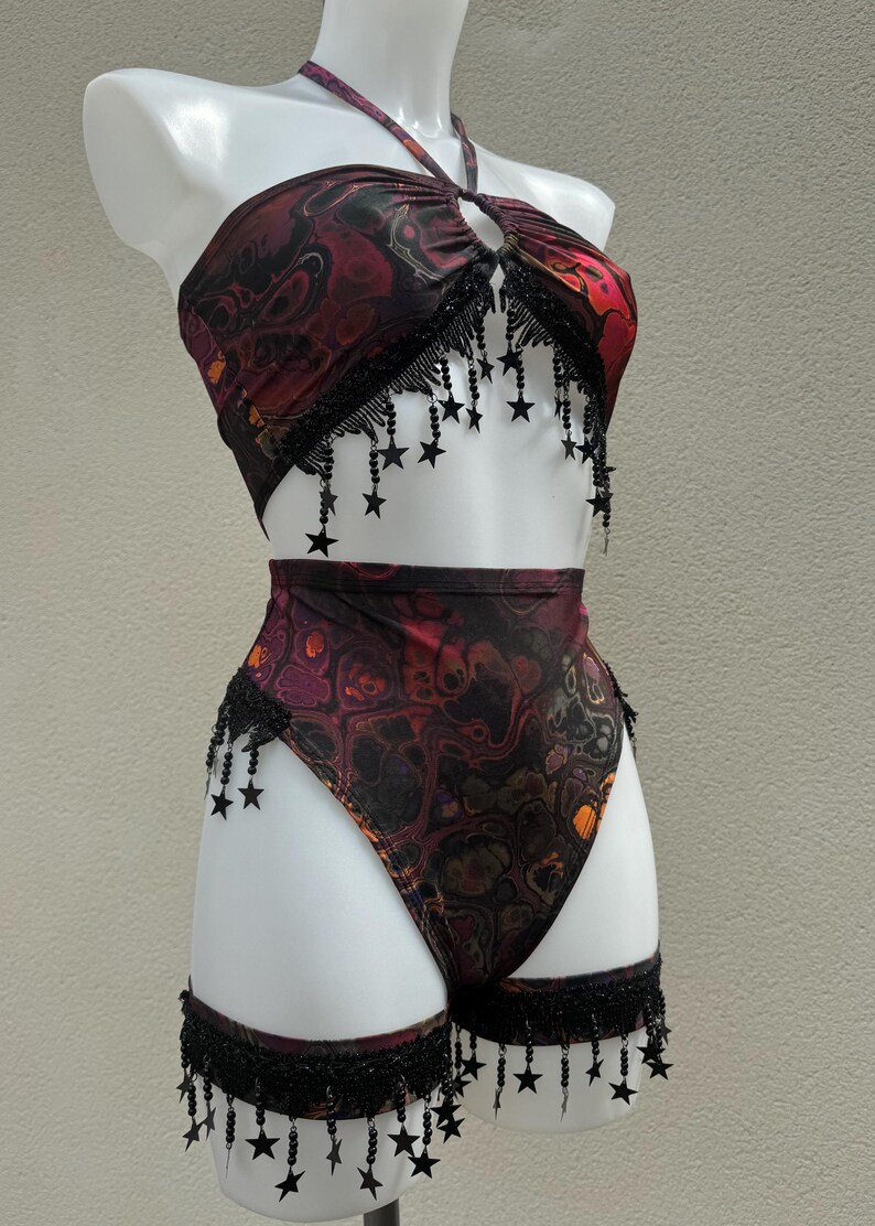 May include: A two-piece outfit featuring a halter top and high-waisted bottoms. The fabric displays a swirling pattern in red, black, and orange. Black star-shaped fringe decorates the top and bottom edges, adding a decorative element.