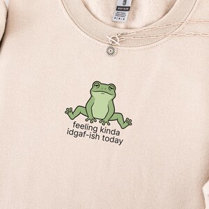 May include: Sand-colored sweatshirt with a green frog graphic and the text "feeling kinda idgaf-ish today." The sweatshirt is paired with blue jeans and a silver necklace. The design is casual and humorous.