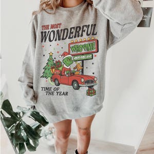 May include: A gray sweatshirt featuring a graphic of the Grinch driving a red car with a dog, a Christmas tree, and the text "The Most Wonderful Time of the Year." The design includes the word "Whoville."