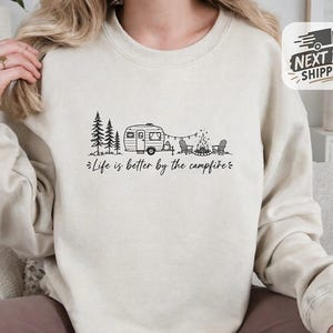 May include: A beige crewneck sweatshirt featuring a black line art graphic of a camping trailer, pine trees, two chairs, and a campfire. Below the illustration, the text reads, "Life is better by the campfire" in a cursive script font.