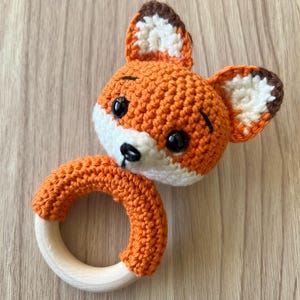 Op de afbeelding: Een handgemaakte gehaakte baby rammelaar vos. De vos heeft een oranje kop met witte en bruine accenten, zwarte ogen en een witte snuit. De rammelaar heeft een houten ring basis, bedekt met oranje haakwerk.