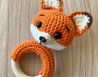 Op de afbeelding: Een handgemaakte gehaakte baby rammelaar vos. De vos heeft een oranje kop met witte en bruine accenten, zwarte ogen en een witte snuit. De rammelaar heeft een houten ring basis, bedekt met oranje haakwerk.