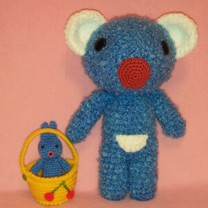 Pode incluir: Dois animais de pelúcia de crochê, um urso azul e um coelho azul, em um fundo rosa. O urso tem um nariz vermelho e orelhas brancas. O coelho está sentado em uma cesta amarela com cerejas vermelhas.