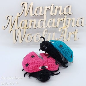 Puede incluir: Dos juguetes de mariquita de crochet, uno rosa y otro turquesa, con patas y cabezas negras. Los juguetes de mariquita están sentados sobre una superficie blanca con un letrero de madera detrás que dice "Marina Mandarina Woolly Art".