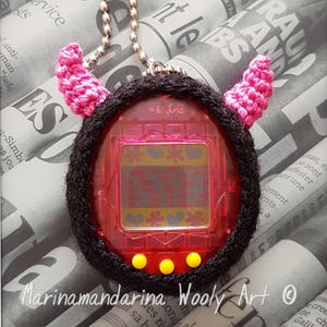 Puede incluir: Un llavero Tamagotchi de crochet rosa y negro con cuernos rosas. El Tamagotchi es rojo y tiene una pantalla con un patrón de flores y corazones. El llavero tiene una cadena plateada.