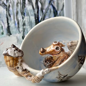 Peut inclure: Un bol en céramique fantaisiste avec un intérieur et un extérieur bleu clair, représentant une créature brune et beige tenant un cupcake. Le bol a une scène d'hiver peinte à l'extérieur, posé sur une surface blanche.