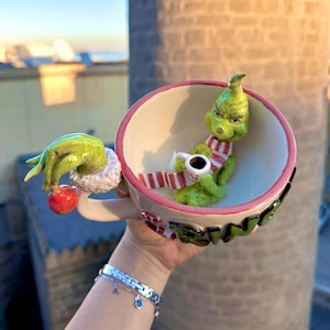 Könnte beinhalten: Eine Keramiktasse mit einer grünen Grinch-Figur im Inneren, die einen rot-weiß gestreiften Schal trägt und eine kleine weiße Tasse hält. Der Griff ist eine grüne Hand, die eine rote Verzierung hält. Die Tasse hat einen rosa Rand und grüne Schrift.
