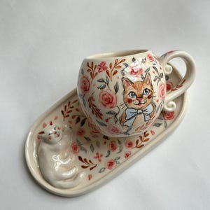 Può includere: Un set di tazza e piattino in ceramica. La tazza e il piattino sono color crema e decorati con rose rosa, foglie verdi e un gatto dei cartoni animati con un papillon blu. Il piattino presenta una figura di gatto 3D.