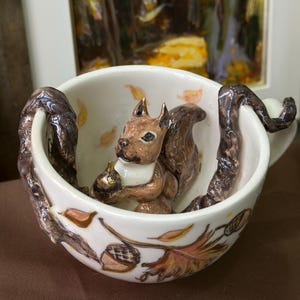 Puede incluir: Una taza de té de cerámica blanca con una figura de ardilla marrón en el interior, sosteniendo una bellota. La taza tiene asas en forma de rama marrón y está decorada con hojas y bellotas de otoño.