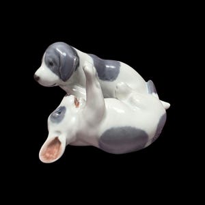 Puede incluir: Figura de cerámica con dos cachorros jugando. Uno está tumbado boca arriba, el otro de pie encima. Los cachorros son blancos con manchas grises y detalles, con un acabado brillante. Una pieza decorativa encantadora.