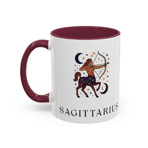 May include: A white ceramic mug with a dark maroon handle and interior rim. The front features a graphic illustration of a Sagittarius centaur archer surrounded by stars, moons, and constellations, with the word SAGITTARIUS printed below.