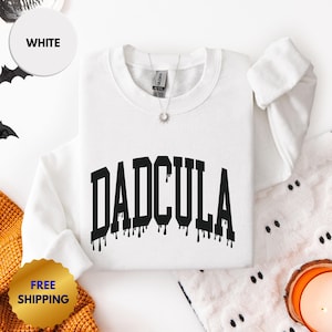 May include: A white long-sleeve crewneck sweatshirt featuring the word DADCULA printed in a black, dripping, varsity-style font across the chest. The garment has a ribbed collar, cuffs, and hem, with a silver-toned sunburst pendant necklace.