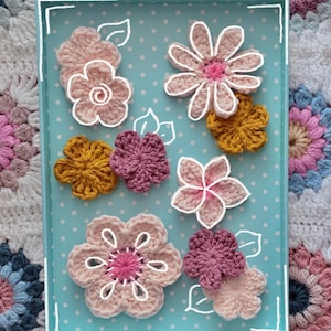 May include: A collection of handmade crochet flowers in various colors, including pink, yellow, and purple, arranged on a light blue tray with white polka dots. The flowers have white outlines and are set against a floral background.