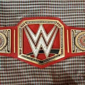 May include: A red wrestling championship belt with gold and silver accents. The center plate features a large "W" logo, with "UNIVERSAL CHAMPION" text below. Two side plates display the WWE logo. The belt is made of red leather.