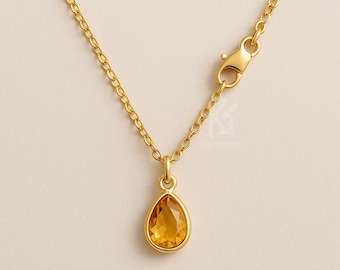 Könnte beinhalten: Eine goldfarbene Halskette mit einem tropfenförmigen Anhänger. Der Anhänger hat einen gelb-orangenen Edelstein, der in einer goldenen Lünette gefasst ist. Kette und Verschluss sind ebenfalls goldfarben, was ein stimmiges Design ergibt. Ein zarter Schmuck.